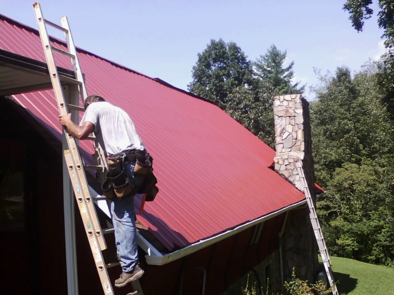 Roofer on a ladder beside a red metal roof and stone chimney — Soffit & Fascia Repair by Roofing Brothers in Mableton